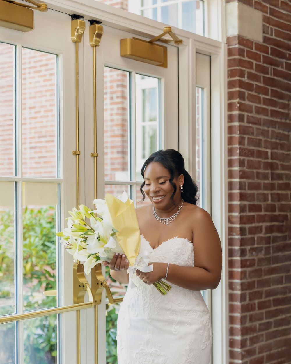 Smiling bride in strapless lace gown holding white floral bouquet near gold-framed glass doors with brick wall backdrop.