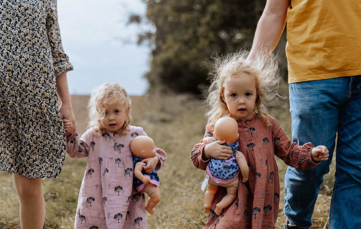 Children in pink dresses walk along a path while holding dolls and their parents' hands on a sunny day.