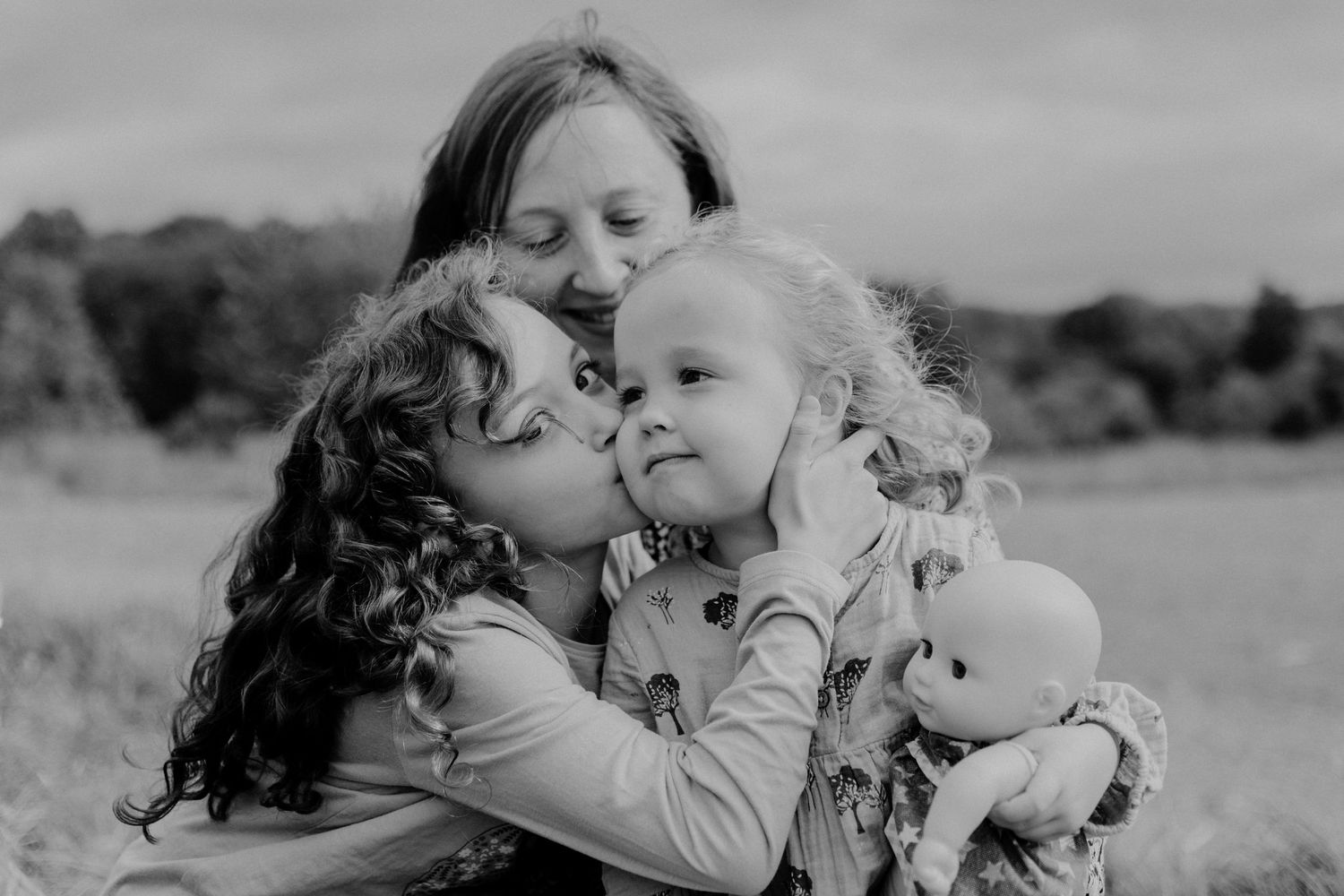 A sweet black and white photograph of two people sharing a tender kiss with a toddler while holding a doll outdoors.