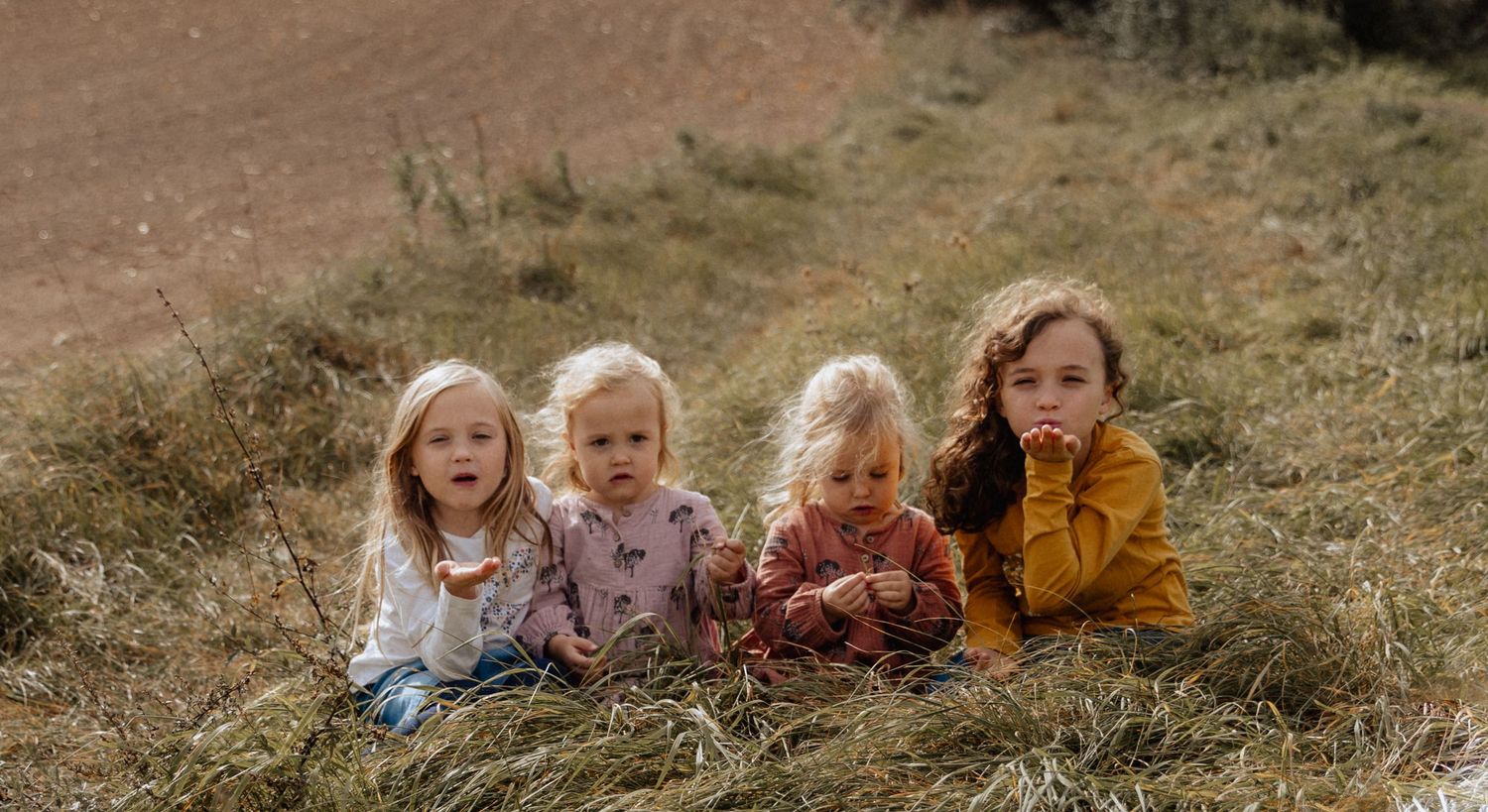 Four children in cozy sweaters lie together on a grassy hillside in autumn.