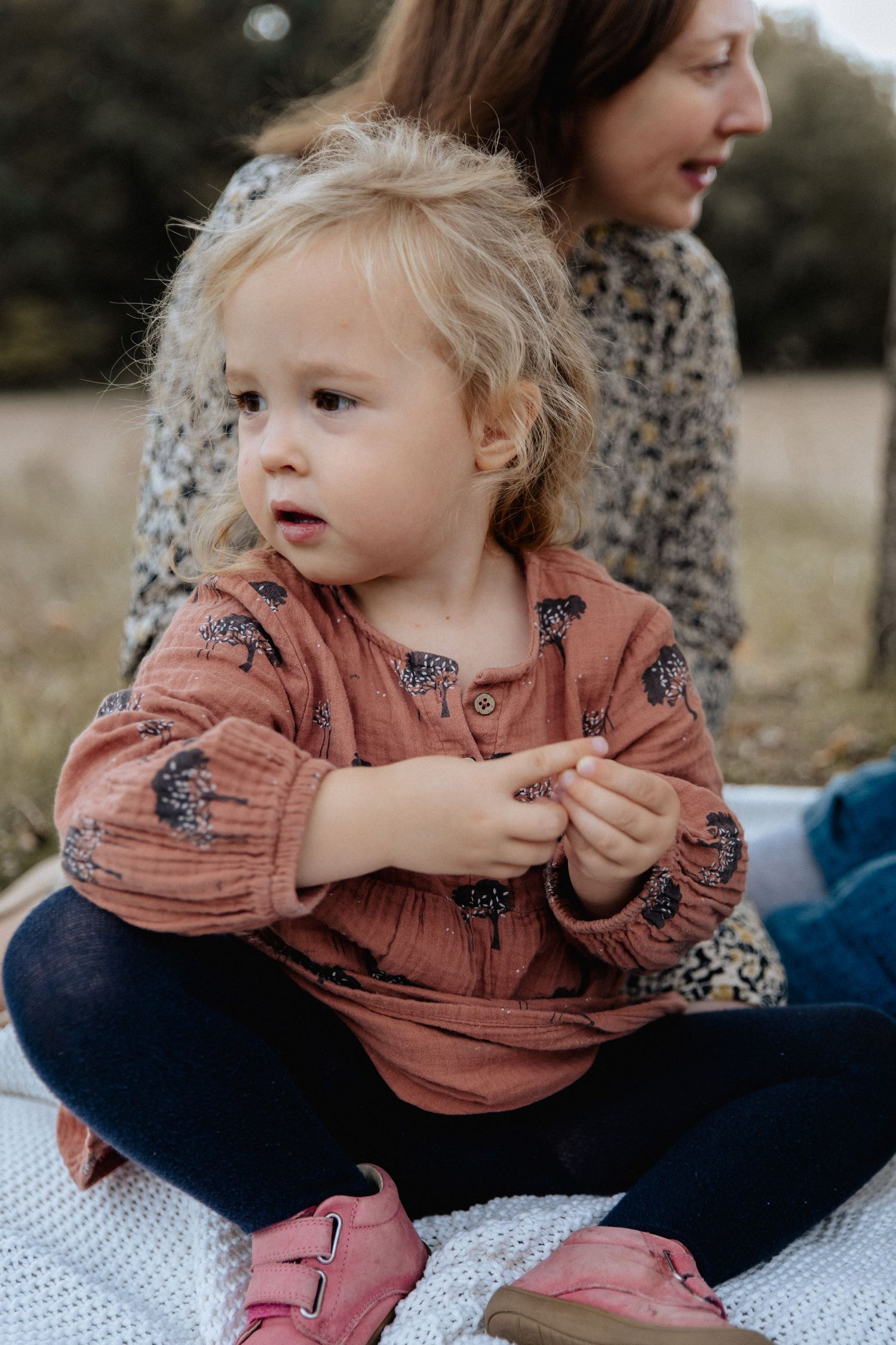 A child in a salmon-colored patterned blouse sits on a blanket outdoors with pink shoes and navy leggings.