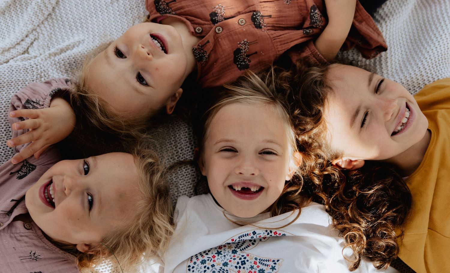 Four children lying on the floor in a circle formation with joyful smiles and laughter, photographed from above.