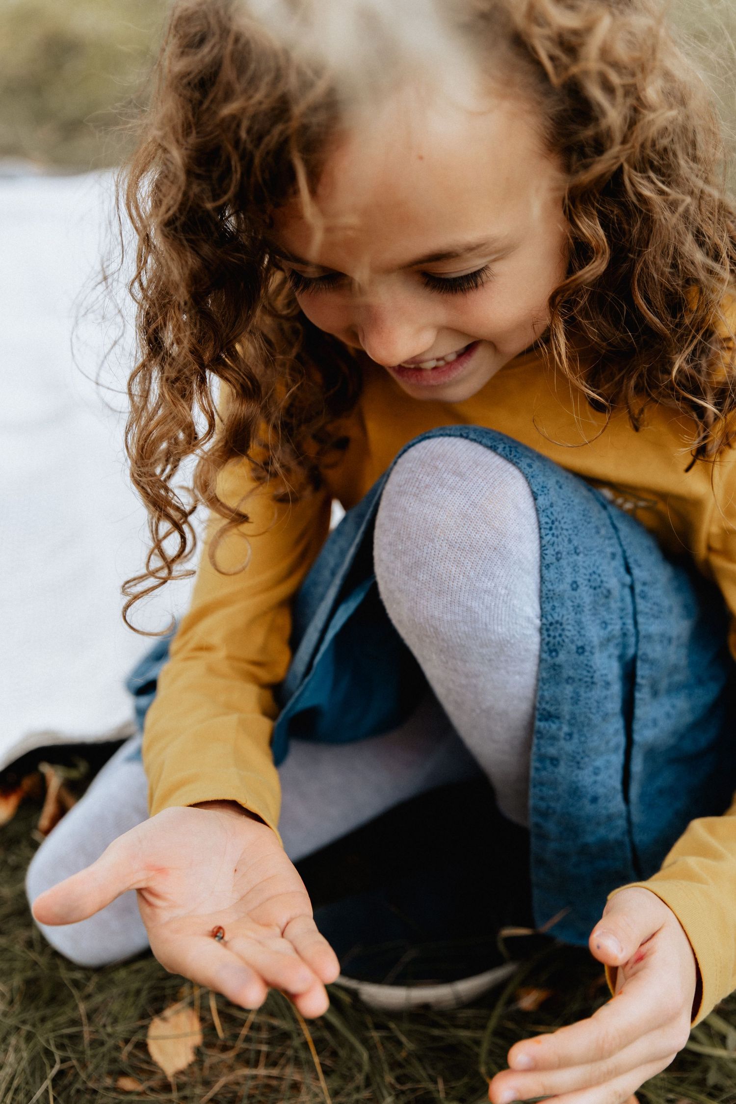 Young person in a mustard yellow sweater and denim smiles while crouching down to look at something on the ground.