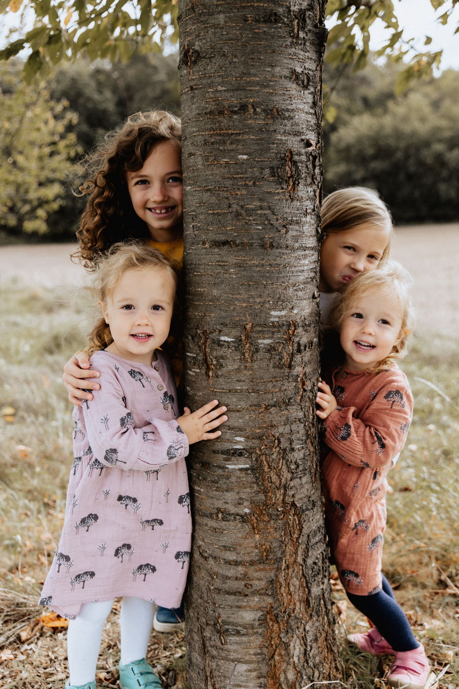 Children peek playfully around a large tree trunk in a peaceful outdoor autumn setting.