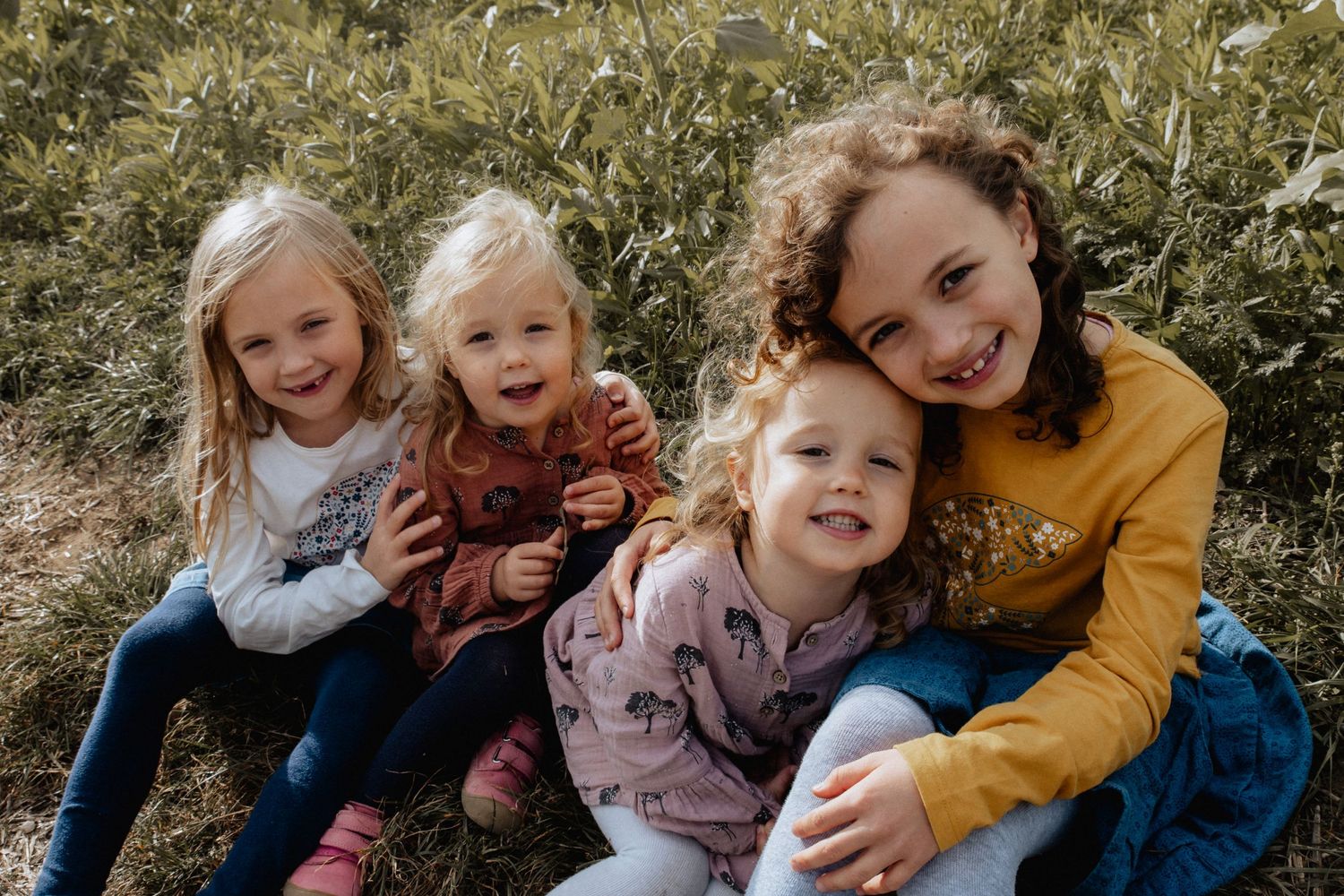 Four children sitting together outdoors in a field laughing and smiling on a sunny day.
