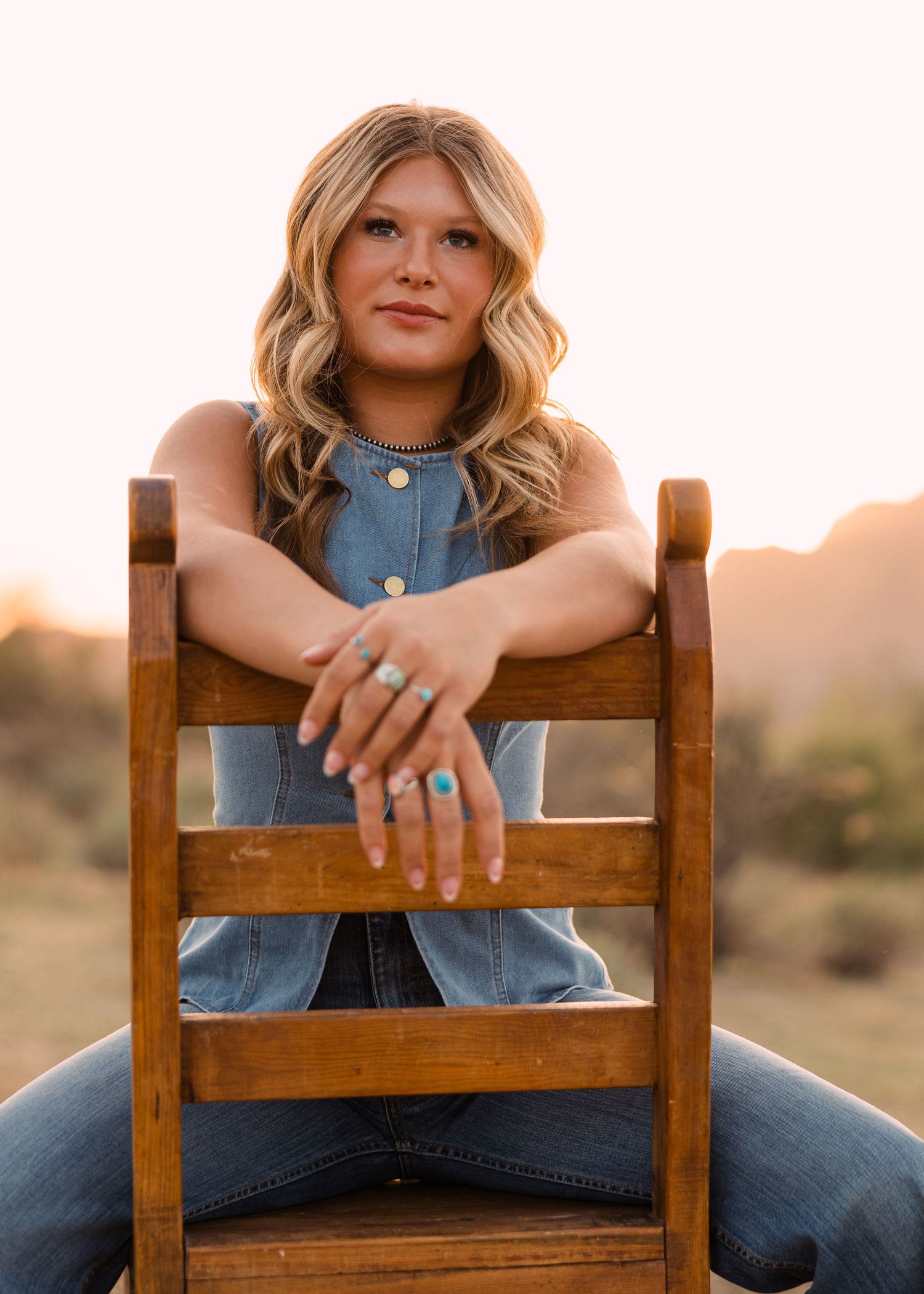 A young woman with wavy blonde hair and turquoise rings sits on a wooden chair in a desert landscape at sunset.