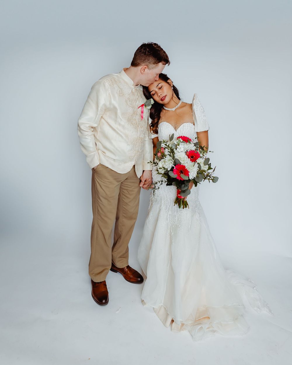 A romantic wedding couple poses against a white backdrop with the bride holding a red and white bouquet.