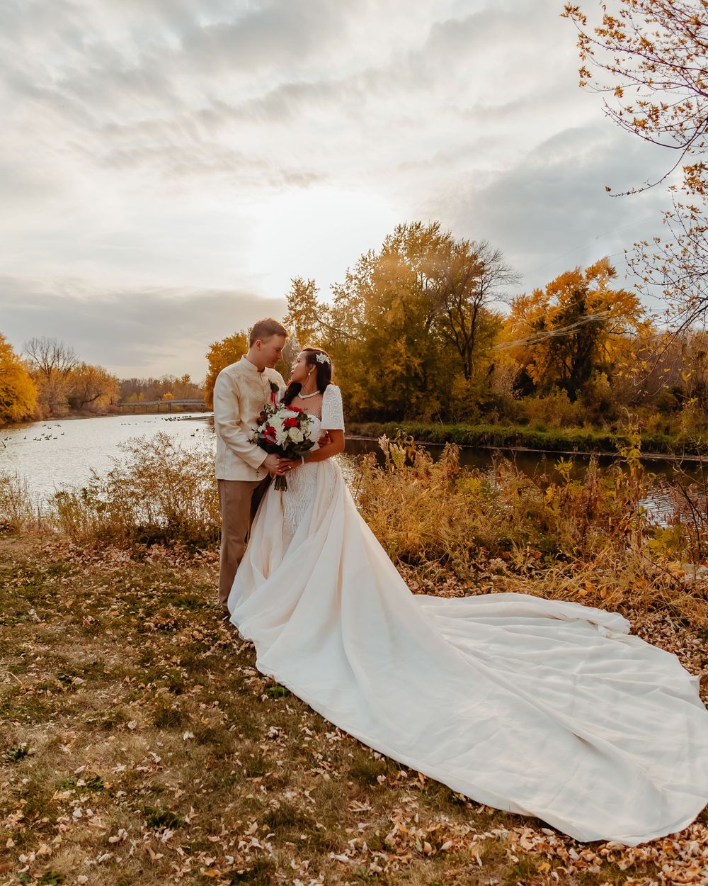 A beautiful autumn wedding photo by a lake with golden trees in the background.