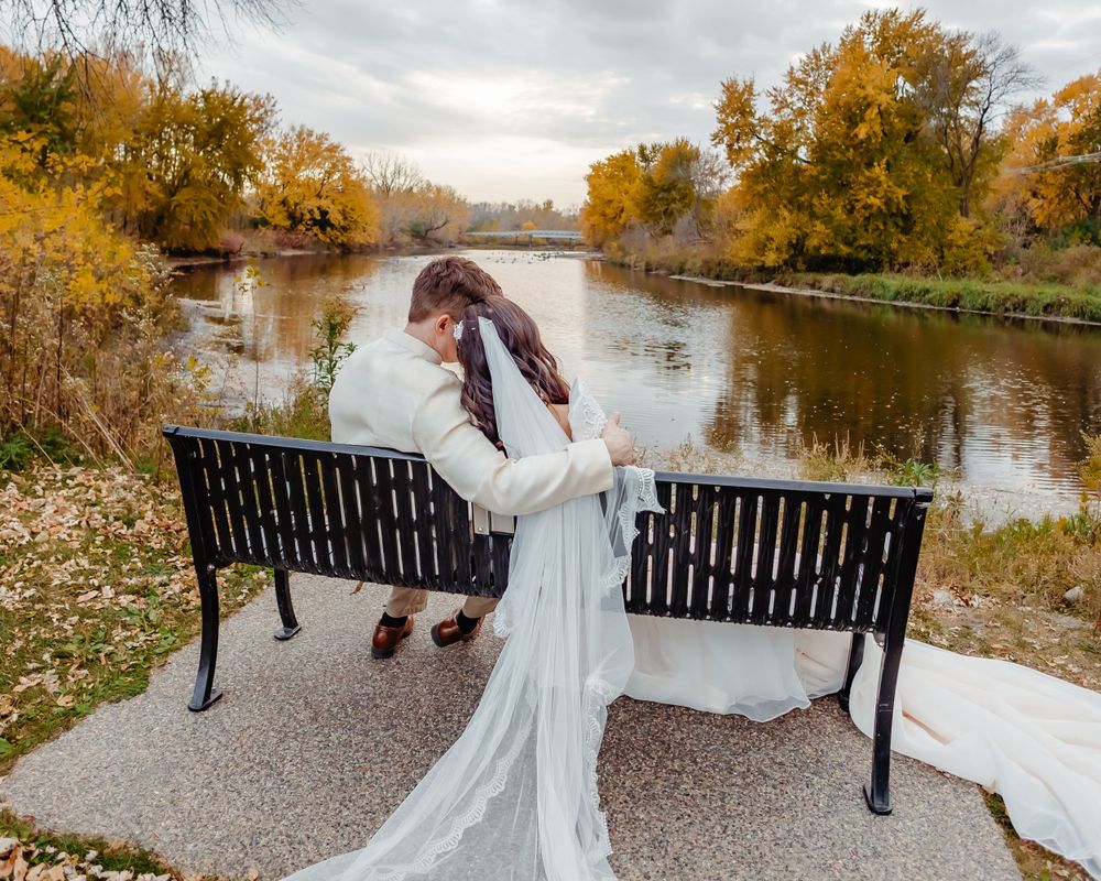 A romantic wedding moment captured on a park bench overlooking an autumn lake.