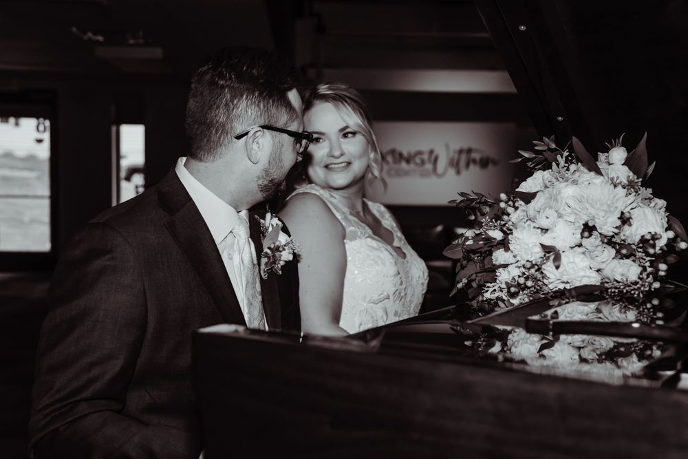 A black and white wedding photo of a couple at a piano with elegant floral arrangements.