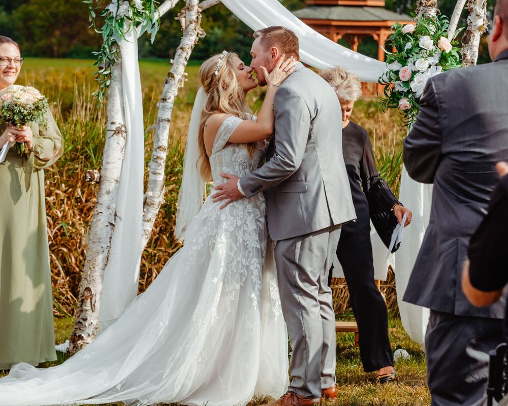 Wedding kiss under white arch decorated with greenery during outdoor fall ceremony.