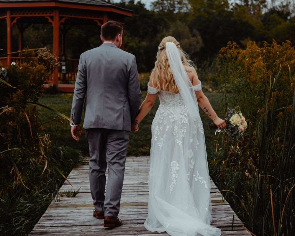 A couple walks along a wooden boardwalk surrounded by wildflowers with a gazebo in the background.