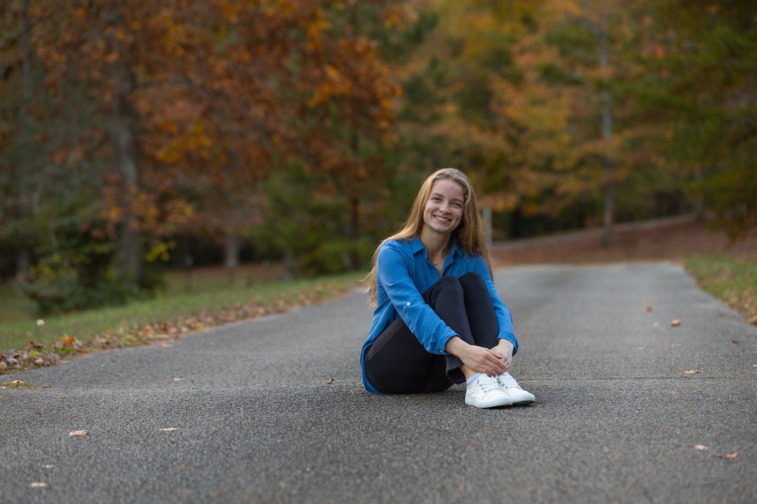 A person in a blue sweater sits on a paved path surrounded by autumn foliage.