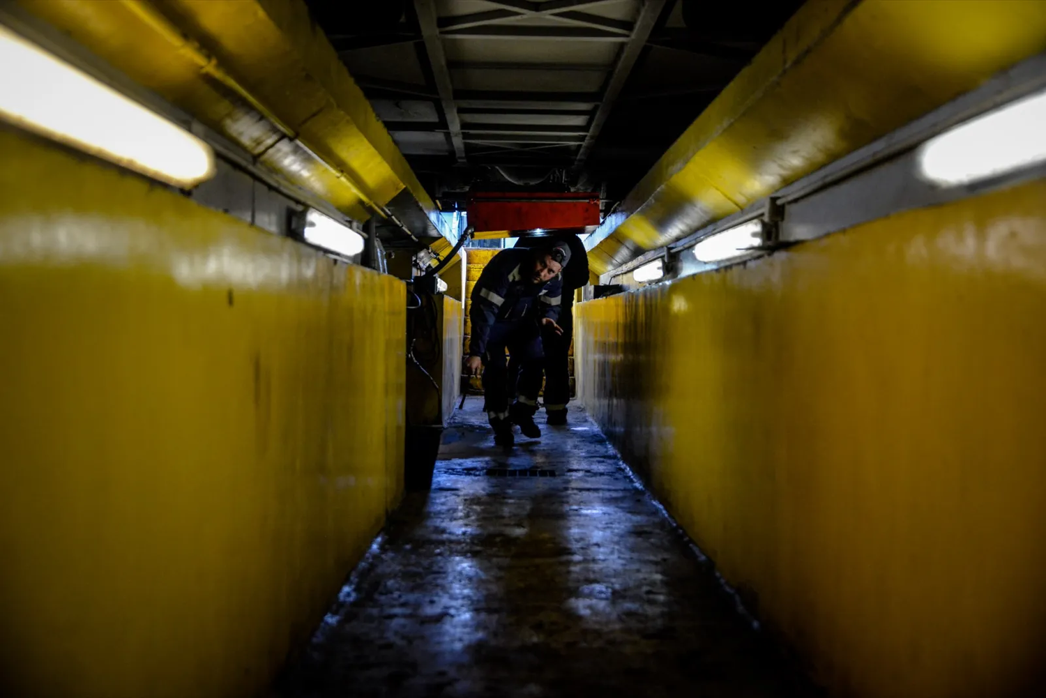 Couloir industriel jaune avec silhouettes humaines en fond, ambiance souterraine.