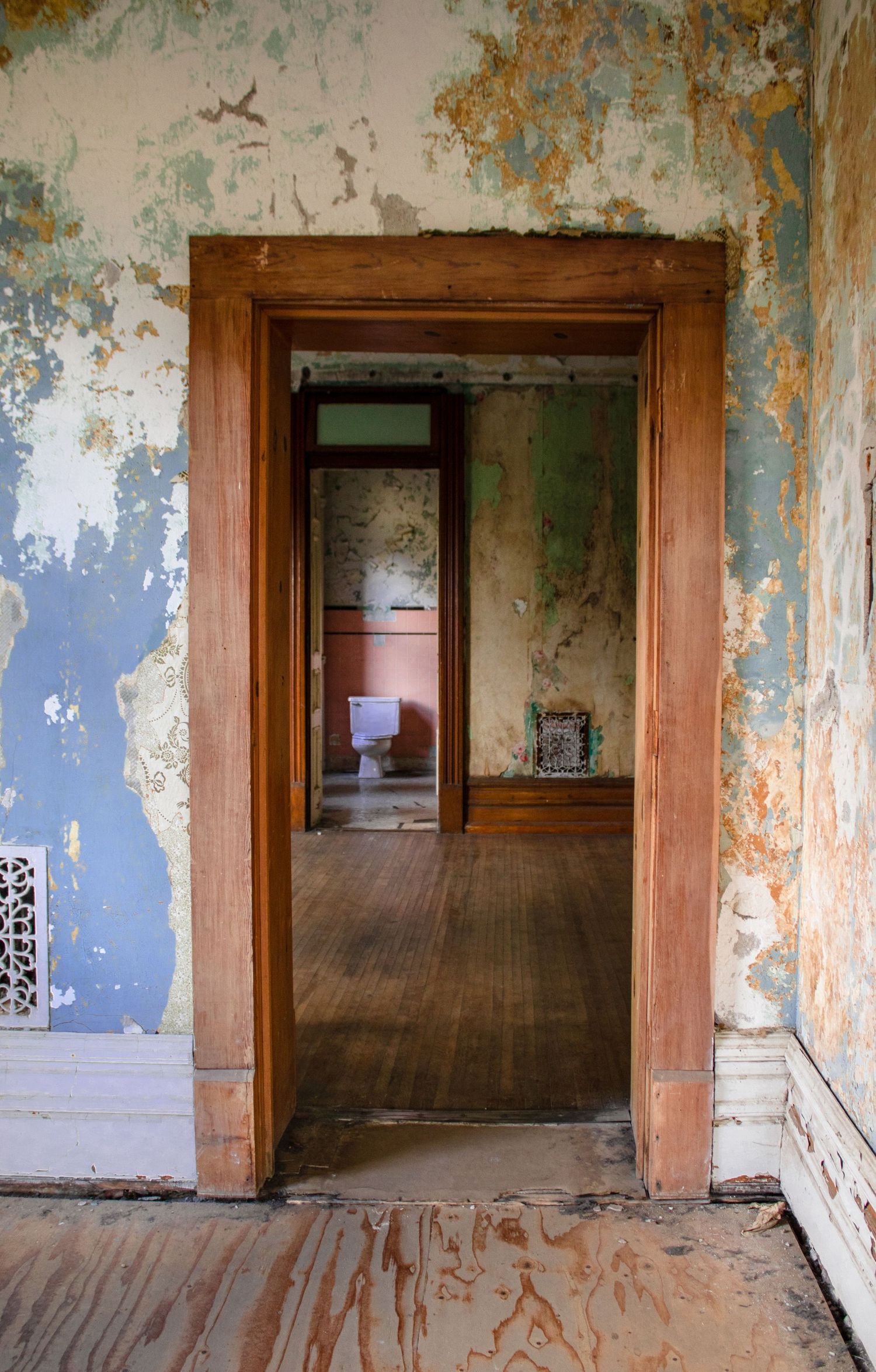 Abandoned room with peeling paint walls and wooden doorframe revealing toilet in distance.