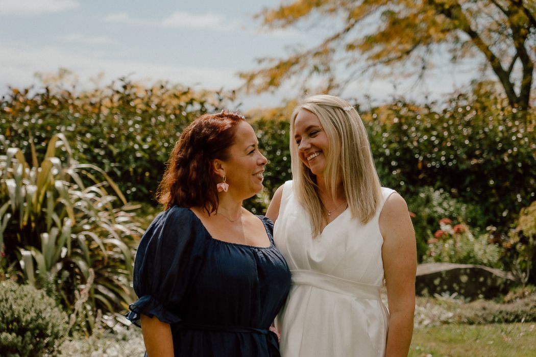 Two women in a garden, one in navy blue dress and one in white bridal gown, smiling at each other.