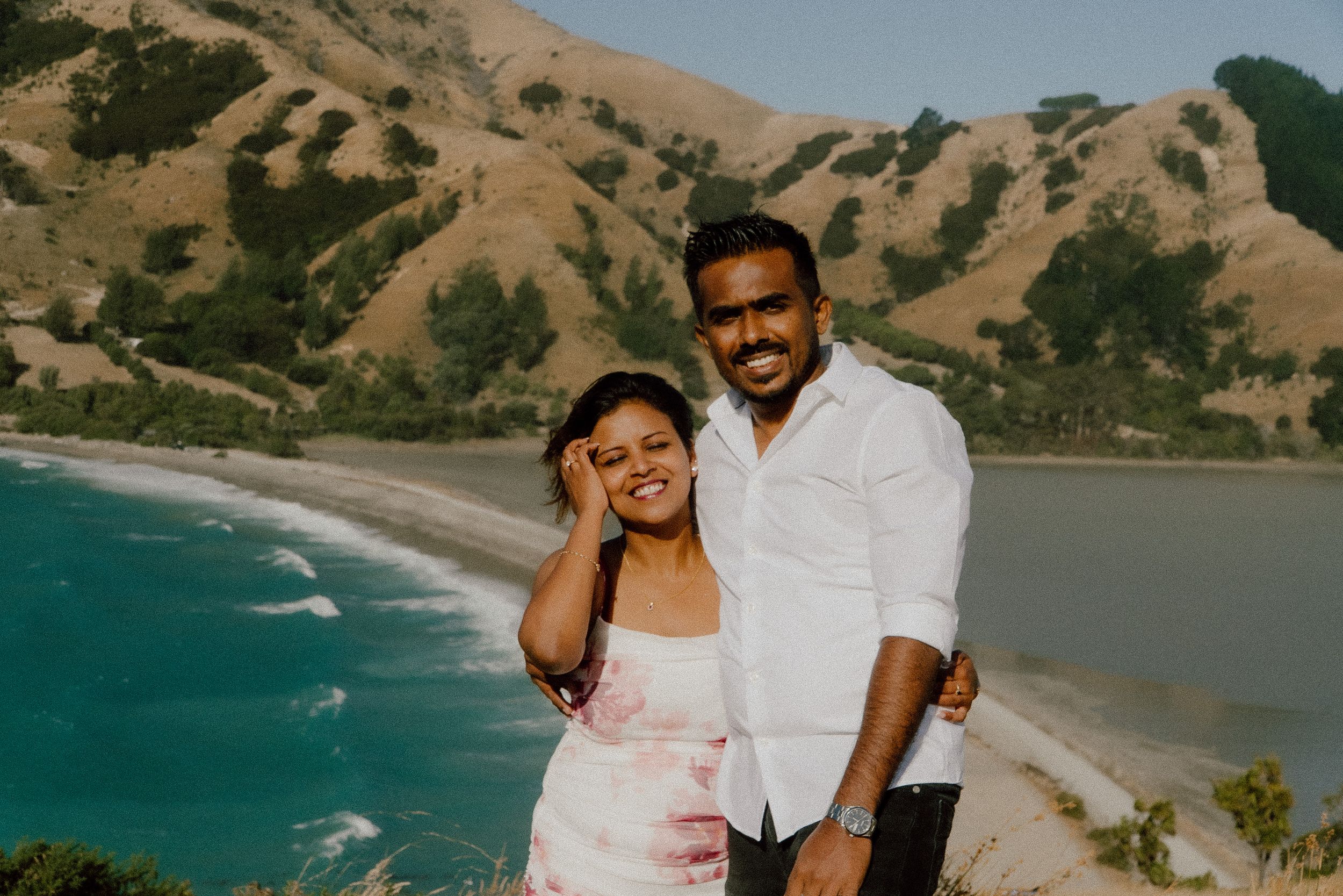 Smiling couple embracing on a hilltop overlooking a stunning turquoise bay and green hills.