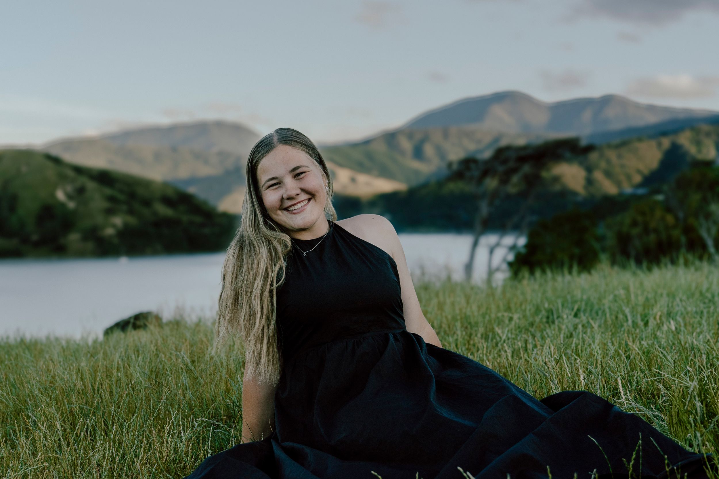 A woman in a black dress reclines on grassy hillside overlooking a scenic mountain lake view.