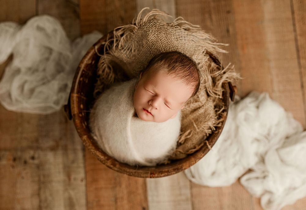 Baby Easton swaddled in cream, peacefully sleeping in a wooden bucket on rustic wood flooring at Bailey's Photography in Minford, Ohio