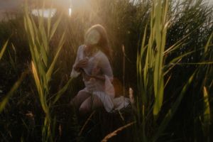 Woman in white dress kneeling among tall green reeds bathed in warm golden sunset light.