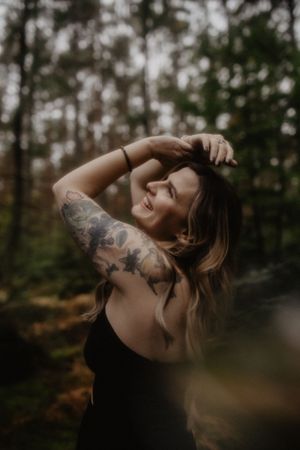 Smiling woman with floral arm tattoos posing in a forest, wearing a black top, with long blonde hair.