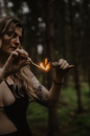 Tattooed woman lighting a sage bundle with a match in a dark moody forest setting.