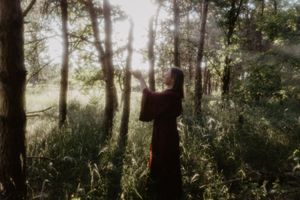 Woman in dark dress reaching toward sunlight filtering through trees in a misty forest.