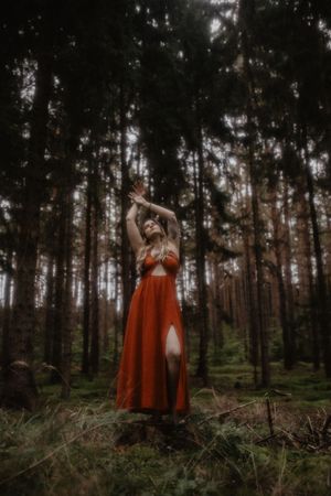 Woman in red dress standing on a stump in a dark forest with arms raised gracefully upward.