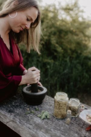 Woman in red blouse grinding herbs with mortar and pestle on rustic wooden table with glass jars outdoors.