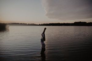 Person standing in calm lake at dusk with arms raised, misty water and dramatic cloudy sky in background.