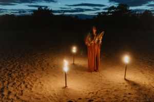 Robed figure stands in a circle of glowing candles on sandy ground at twilight.
