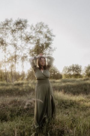 Woman in olive green maxi dress poses gracefully in a sunlit meadow with trees in the background.