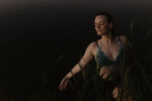 Woman in teal bikini top sits peacefully among tall grass near dark water with eyes closed at dusk.