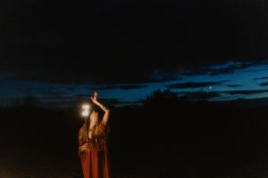 Figure in red robes raises torch against dark evening sky.
