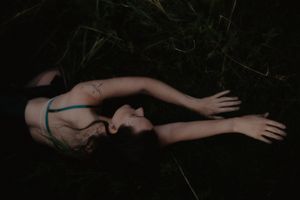A woman in a bikini top lies among dark green grass and reeds in a moody, atmospheric outdoor setting.