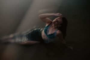 A woman in a teal outfit reclines dramatically on a dark floor with moody atmospheric lighting.