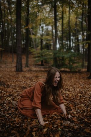 Person in rustic dress crouches among autumn leaves in moody forest setting.