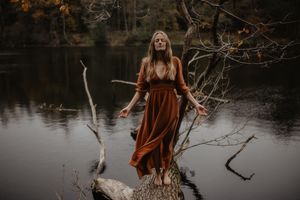 Figure in flowing rust-colored dress stands on fallen log over still water.