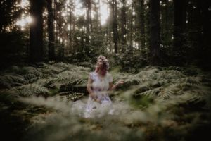 A woman in a white dress sits peacefully among lush green ferns in a misty forest with sunlight filtering through trees.