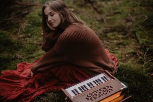 Person in brown sweater sits with harmonium among moss and red fabric outdoors.