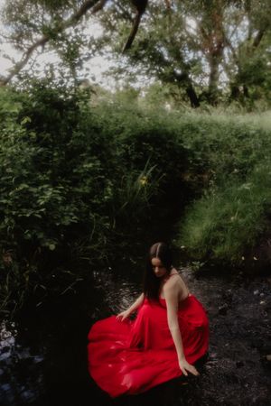 A red dress creates a striking contrast against dark water and lush greenery in a forest stream.