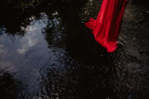 The hem of a red dress reflects in dark water, creating an artistic composition.