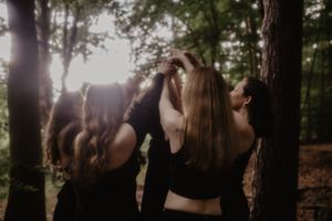 Four women in black outfits gather among sunlit forest trees, their long hair flowing as they reach upward together.
