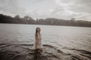 Woman in white dress emerging from a misty lake surrounded by trees under a cloudy sky.