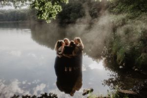 Group of friends embracing by a misty lake surrounded by lush green trees reflected in calm water.