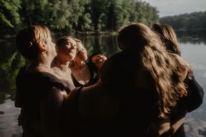 Group of women embracing near a tranquil lake surrounded by lush green trees in soft natural light.