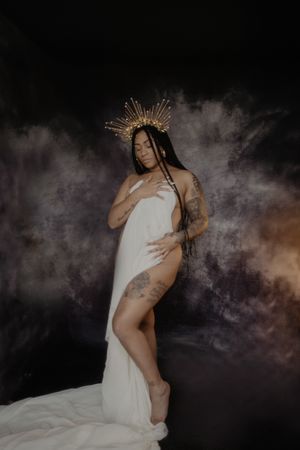 Artistic portrait in white fabric with a gold sunburst crown against a dark smoky backdrop.