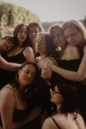 Seven women in black dresses embrace closely together outdoors, creating an intimate and artistic group portrait.