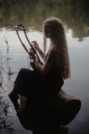 Silhouette sitting by water playing stringed instrument in moody evening light.