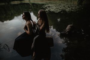 Two figures in dark dresses sitting on log by tranquil water.